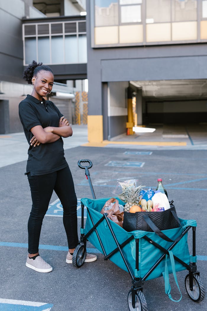 services-01 Black woman posing with a grocery cart filled with items in an outdoor parking area.